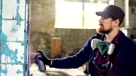 Serious-man-is-painting-graffiti-on-dirty-pillar-in-old-empty-house-with-aerosol-paint.-Guy-is-wearing-casual-clothes-and-trendy-cap,-he-has-protective-gloves-and-mask.