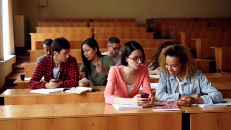 Junge-Menschen-nutzen-Smartphones-und-sitzen-am-Schreibtisch-im-Hörsaal-an-der-Universität-zu-sprechen.-Geräumiges-Zimmer-mit-Holzmöbeln,-Frauen-und-Männer,-sind-Notebooks-sichtbar.