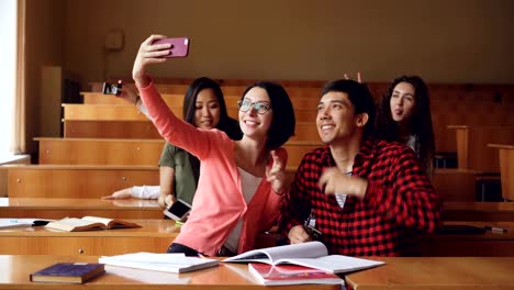 Fröhlichen-Studenten-nehmen-Selfie-im-Hörsaal-sitzen-zusammen-an-Schreibtischen-und-halten-Smartphones.-Moderne-Technologie,-Selbstporträt-und-Bildung-Konzept.