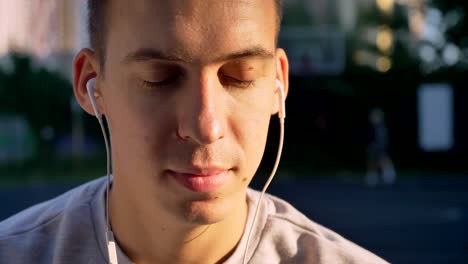 Retrato-de-joven-guapo-hombre-escuchar-música-mediante-auriculares-y-mirando-a-cámara,-gente-jugando-al-aire-libre-fondo