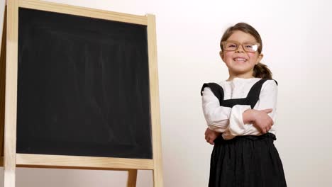 A-happy-girl-dressed-as-a-teacher-in-front-of-a-small-blackboard-holds-her-arms-folded-and-smiles.