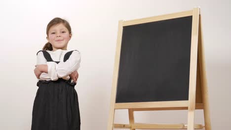 A-happy-girl-dressed-as-a-teacher-in-front-of-a-small-blackboard-holds-her-arms-folded-and-smiles.