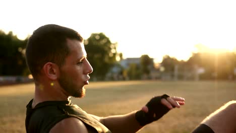 A-young-boxer-in-black-bandages-working-out-outdoors-on-the-stadium---punching.-Kickboxing-training.-Sun-shining-on-the-background