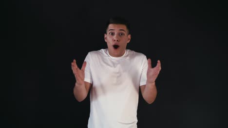 Excited-and-amazed-young-man-in-white-shirt-stands-and-looks-on-camera.-He-lifted-up-his-his-hands-and-express-emotions.-Guy-change-it-to-smile-and-wonder.-Isolated-on-black-background