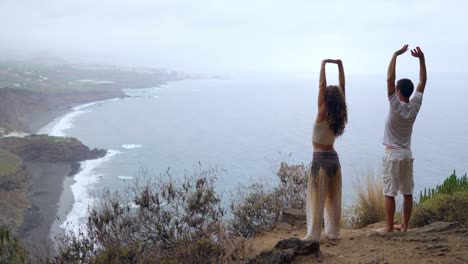 Un-hombre-y-una-mujer-de-pie-en-el-borde-de-un-acantilado-con-vistas-al-océano-levantan-sus-manos-y-aspirar-el-aire-del-mar-durante-yoga