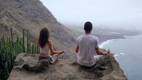 Un-hombre-y-una-mujer-sentada-en-la-cima-de-una-montaña-mirando-el-mar-sentado-en-una-piedra-meditando-en-posición-de-loto.-La-vista-desde-la-parte-posterior.-Islas-Canarias