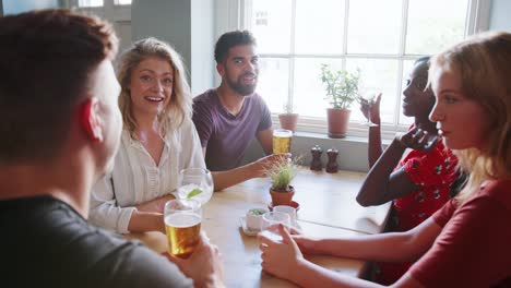 Five-happy-young-adult-friends-sitting-with-drinks-at-a-table-in-a-pub-talking,-close-up