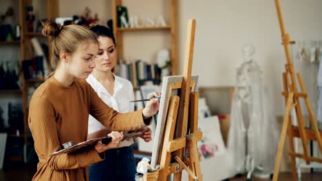 Süße-Teenager-Mädchen-lernt-Malerei-von-erfahrenen-Lehrer-an-der-Kunstschule-in-Arbeitszimmer-mit-Kunstwerken-und-hölzernen-Staffeleien.-Künstler-lehrt-reden-und-auf-Bild-zeigen.