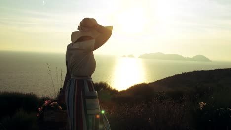 Beautiful-young-woman-wearing-fashion-colorful-dress-with-skirt-and-hat-carrying-flowers-in-basket-at-sunset-on-Ponza-Island-mountain-Italy.