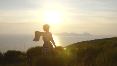 Schöne-junge-Frau-tragen-bunte-Kleid-Mode-mit-Rock-und-Hut-Stand-mit-Blumen-im-Korb-bei-Sonnenuntergang-auf-der-italienischen-Insel-Ponza-Berg-Küste.