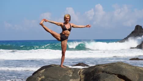 Young-girl-in-bikini-balancing-stands-on-one-leg-doing-yoga-standing-on-a-rock-on-the-ocean-beach-with-black-sand.-Meditation-through-relaxation.-Gymnastics