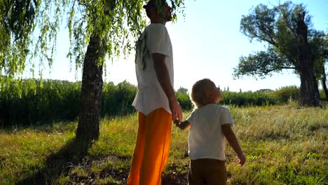 Niño-pequeño-mirando-hacia-arriba-en-un-árbol-alto-durante-la-marcha-con-su-papá-a-través-del-parque-en-día-de-verano.-Joven-padre-y-su-hijo-lindo-pasar-tiempo-juntos-al-aire-libre.-Luz-del-sol-en-el-fondo.-Lenta-de-cerca