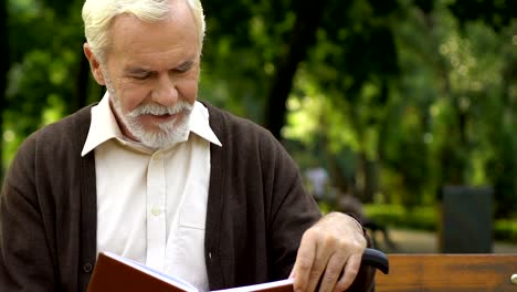 Old-wrinkled-man-with-cane-sitting-on-bench-and-reading-book-at-green-park