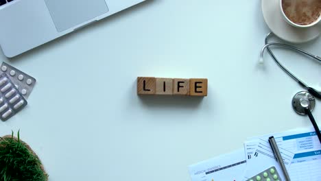 Life-word-made-of-wooden-cubes-on-table,-doctor-hands-showing-toy-heart-top-view