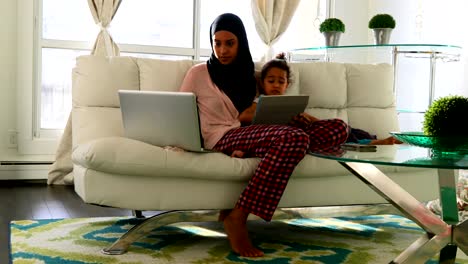 Joven-madre-e-hija-usando-portátil-y-tableta-digital-en-la-sala-de-estar-4k