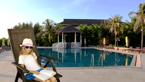 Young-caucasian-girl-sitting-on-armchair-and-reading-book.-Swimming-pool-and-palm-trees-at-background.-Have-same-clip-with-transparent-background-Alpha-Channel.