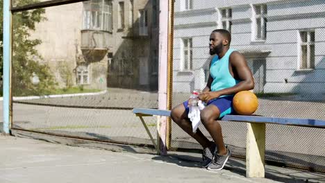 Jugador-de-baloncesto-fuerte-sentado-en-la-banca-y-beber-agua-mineral,-deporte