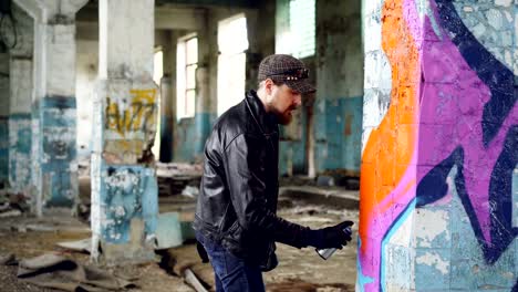 Handsome-bearded-guy-graffiti-artist-is-painting-with-spray-paint-inside-abandoned-building.-Modern-street-art,-youth-subculture-and-creative-people-concept.