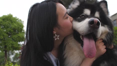 Feliz-joven-mujer-asiática-con-su-perro-malamute-de-Alaska-y-un-beso-al-aire-libre,-4k