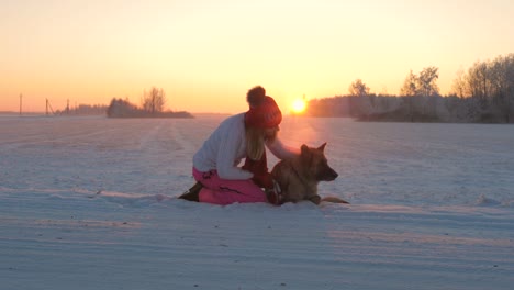 Frau-liebevoll-streicheln-Hundesitting-im-Schnee-an-einem-Winterabend-bei-Sonnenuntergang