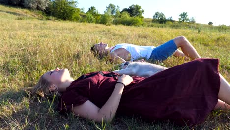 Dolly-shot-of-young-couple-in-love-lying-on-green-grass-at-field-and-stroking-their-siberian-husky-dog-at-sunny-day.-Happy-pair-relaxing-and-enjoying-summer-vacation-at-sunset.-Low-angle-view-Close-up