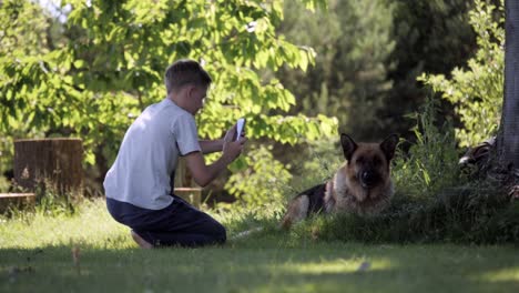 kleiner-Junge-unter-Bild-seines-Hundes-posiert-Deutscher-Schäferhund-mit-einem-Smartphone-in-der-Natur,-grüner-Hintergrund