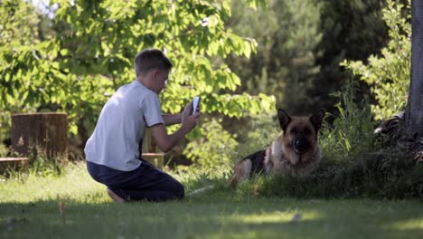 Foto-de-joven-toma-de-su-perro-posando-Pastor-Alemán-con-un-teléfono-inteligente-en-el-fondo-de-la-naturaleza,-verde