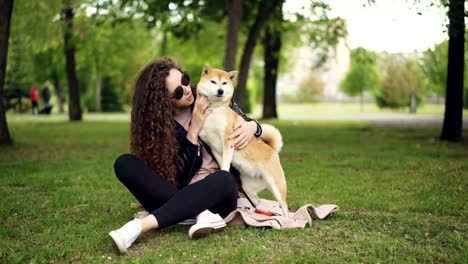 Happy-girl-proud-dog-owner-is-caressing-and-kissing-her-pet-sitting-on-grass-in-the-park-while-the-animal-is-enjoying-love-and-care.-Green-trees-and-lawns-are-visible.