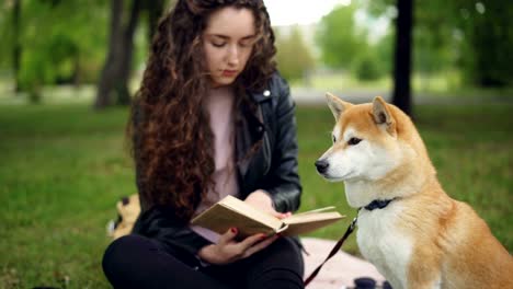 Cheeful-student-is-reading-book-sitting-in-the-park-then-patting-adorable-puppy-and-smiling.-Loving-animals,-intelligent-hobby-and-healthy-lifestyle-concept.