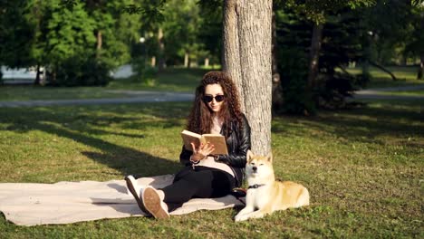 Attraktive-Studentin-liest-Buch-sitzen-auf-Plaid-unter-Baum-im-Stadtpark-mit-ihrem-Hund-liegen-in-der-Nähe-und-Sonnenlicht-zu-genießen.-Hobby,-Freizeit-und-Tiere-Konzept.