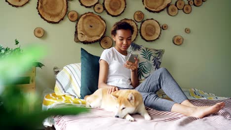 Pretty-young-woman-is-using-smartphone-and-caressing-lovely-puppy-lying-on-bed-near-its-owner-in-modern-style-bedroom-at-home.-Technology,-animals-and-people-concept.