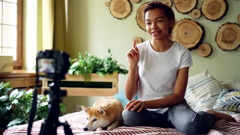 Emocional-chica-blogger-exitoso-está-grabando-video-para-su-vlog-online-sentado-en-la-cama-en-el-apartamento-y-acariciando-lindo-perro-acostado-en-cama.-Concepto-de-blogs-y-los-animales.