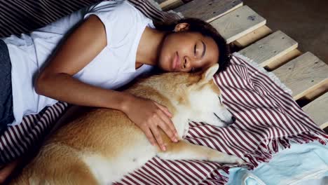 Beautiful-African-American-teenager-and-adorable-pet-dog-are-sleeping-together-on-wooden-bed,-girl-is-wearing-comfortable-pajamas-and-hugging-animal.