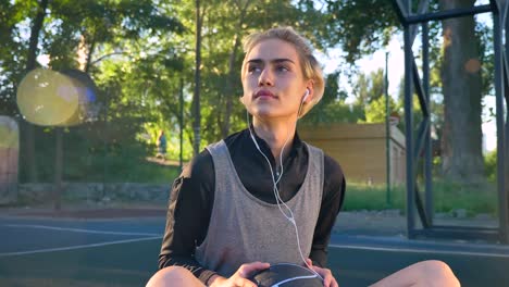 Beautiful-young-woman-sitting-on-basketball-court-and-listening-music-through-earphones,-holding-ball,-open-air-and-lens-flare