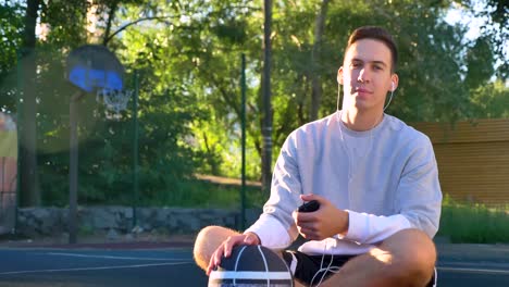 Seguros-de-hombre-joven-sentado-en-la-cancha-de-básquet-y-escuchar-música,-mirando-a-cámara,-sosteniendo-el-teléfono-y-la-bola-del-parque-en-el-fondo,