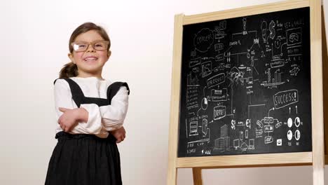 A-happy-girl-dressed-as-a-teacher-in-front-of-a-small-blackboard-holds-her-arms-folded-and-smiles.