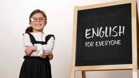 A-happy-girl-dressed-as-a-teacher-in-front-of-a-small-blackboard-holds-her-arms-folded-and-smiles.