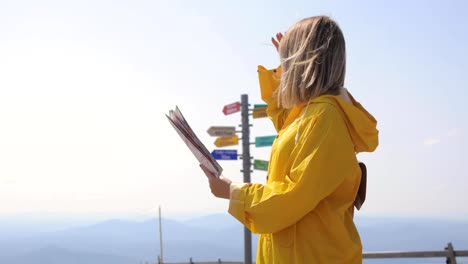 Young-female-hiking-im-yellow-raincoat-with-a-backpack-in-mountains-holding-paper-map-in-hands