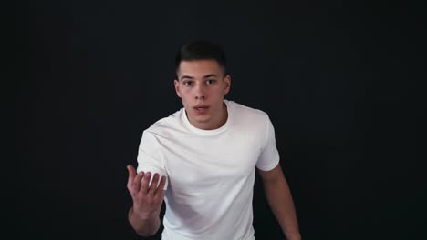 Frustrated-young-man-stands-and-looks-at-camera.-He-doesn't-know-what-to-do.-Guy-tries-to-explain-and-to-reach.-Isolated-on-black-background