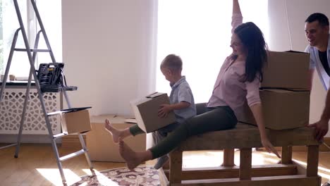 happy-relocation,-young-man-carries-wife-with-little-son-on-table-in-new-apartment-with-of-boxes-with-things