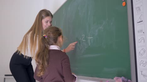 colegiala-con-trozo-de-tiza-escribe-un-ejemplo-en-la-pizarra-con-ayuda-de-una-maestra-en-la-lección-de-matemáticas