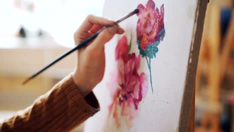 Close-up-shot-of-female-hand-holding-paintbrush-and-painting-bright-flowers-on-canvas-in-light-studio.-Pictorial-art,-artistic-work-and-people-concept.