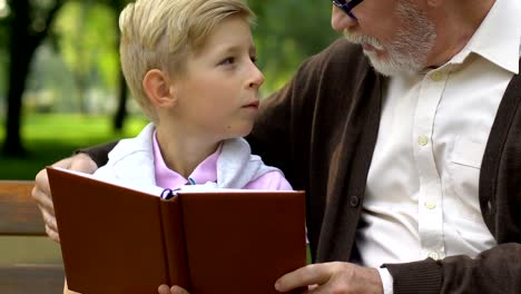 Little-boy-and-grandfather-reading-book,-relaxing-on-bench-in-park,-education