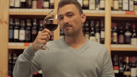 Retrato-de-hombre-guapo-que-beber-vino-tinto.-Hombre-sacude-su-bebida,-levanta-la-Copa-y-toma-un-sorbo-de-vino-con-placer.-El-chico-con-la-Copa-de-vino-en-el-fondo-de-los-estantes-con-botellas-de-vino.