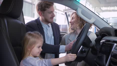 Familienauto-kaufen,-kleines-Kindermädchen-hinter-dem-Steuer-des-neuen-Autos-zusammen-mit-den-Eltern,-während-Kauf-Auto-im-Showroom-Nahaufnahme