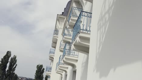 Side-view-of-balconies-with-blue-wrought-iron-railing-of-modern-building,-architectural-background.-Action.-White-wall-of-a-house-with-balconies-against-cloudy-sky