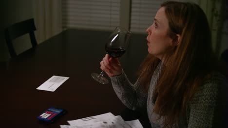 A-woman-looking-over-bills-at-the-table,-looking-stressed-and-drinking-wine