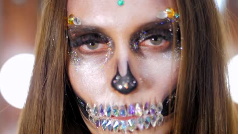 Unheimlich-junges-Mädchen-mit-kreativen-Halloween-Kriegsbemalung-in-der-Umkleidekabine.-Porträt-des-glamourösen-Totenkopf-mit-Strass-und-Pailletten.-Professionelles-Make-up-für-die-Feier.-Slow-motion