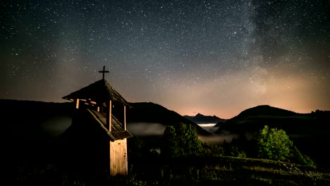 Stars-with-milky-way-galaxy-moving-over-wooden-belfry-in-mountains-and-foggy-valley-rural-country.-Dolly-shot-time-lapse-starry-night-sky