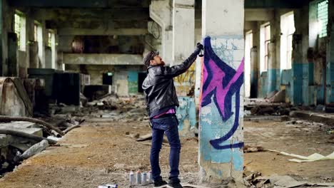 Graffiti-painter-in-casual-clothing-is-drawing-with-spray-paint-on-column-in-spacious-abandoned-building.-Abstract-images,-modern-art,-creative-people-and-hipsters-concept.